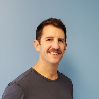 Jason Bodewitz: a man with short dark hair and a dark mustache stands in front of a blue wall, smiling. He is wearing a dark T-shirt.