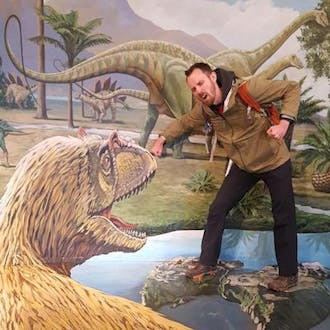 马克·戴恩 pretends his arm is being bitten by a model of a dinosaur. 