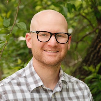 Jason Sobotka's headshot. He wears glasses and a plaid button-up shirt. He smiles, standing in front of trees.