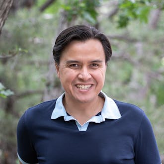 Brian Langevin wears a navy and light blue collared shirt. He is standing in front of trees. He has dark hair and is smiling.