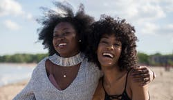 Imagem de duas mulheres jovens se abraçando e sorrindo em direção à câmera em dia ensolarado com vento em seus cabelos.