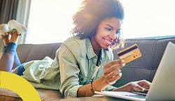 Imagem de mulher jovem deitada em seu sofá com um cartão de crédito em sua mão enquanto olha para seu computador, prestes a realizar a compra de uma passagem online.