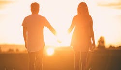 Imagem de um casal composto por um homem e uma mulher, que estão de costas assistindo ao pôr-do-sol. A imagem tem um tom alaranjado pela iluminação do sol.