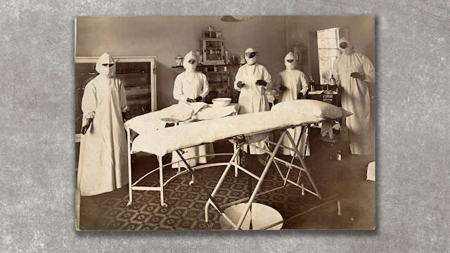 Photograph of an archival black and white photograph resting on a concrete textured background. The archive image shows a group of 5 individuals in an operating theatre, standing around an empty bed. They are all dressed in white gowns with black gloves and full white head marks, which only leave their eyes visible.