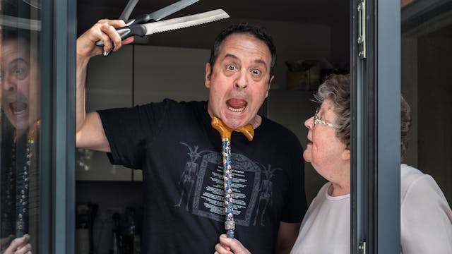 Photograph of a man standing in a glass patio doorway with his mother. The man is looking straight to camera with his mouth open, eyes wide and slightly mad look on his face. In his right hand held above his head are 3 kitchen knives held in a fan shape. His mother is standing to his left in profile, looking up at his face. In her right hand she is holding a walking stick, the handle of which she has tucked under his chin in a disciplinary fashion.