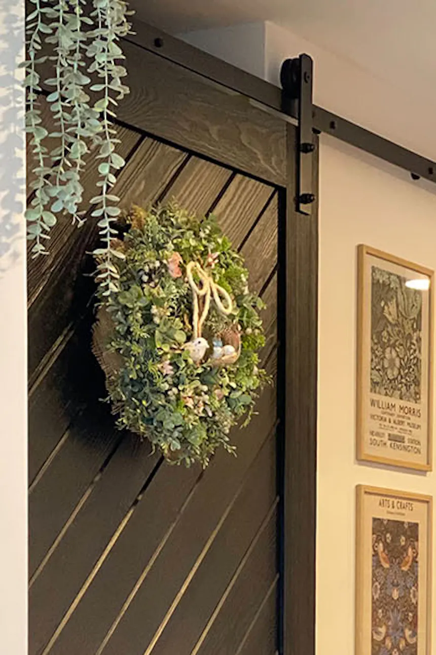 Sliding door for pantry, black solid wood sliding door, decorative wreath on door, red brick on wall, decorative figurines and candle stands on dresser