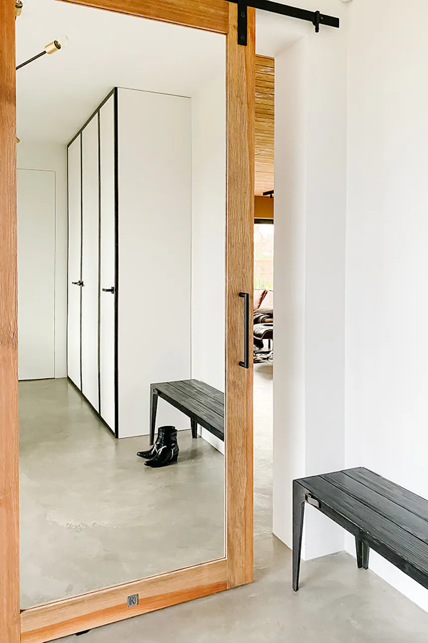 Mirror door in entryway, large mirror in wooden frame, sliding mirror, white walls, black minimalist bench, microcement floor