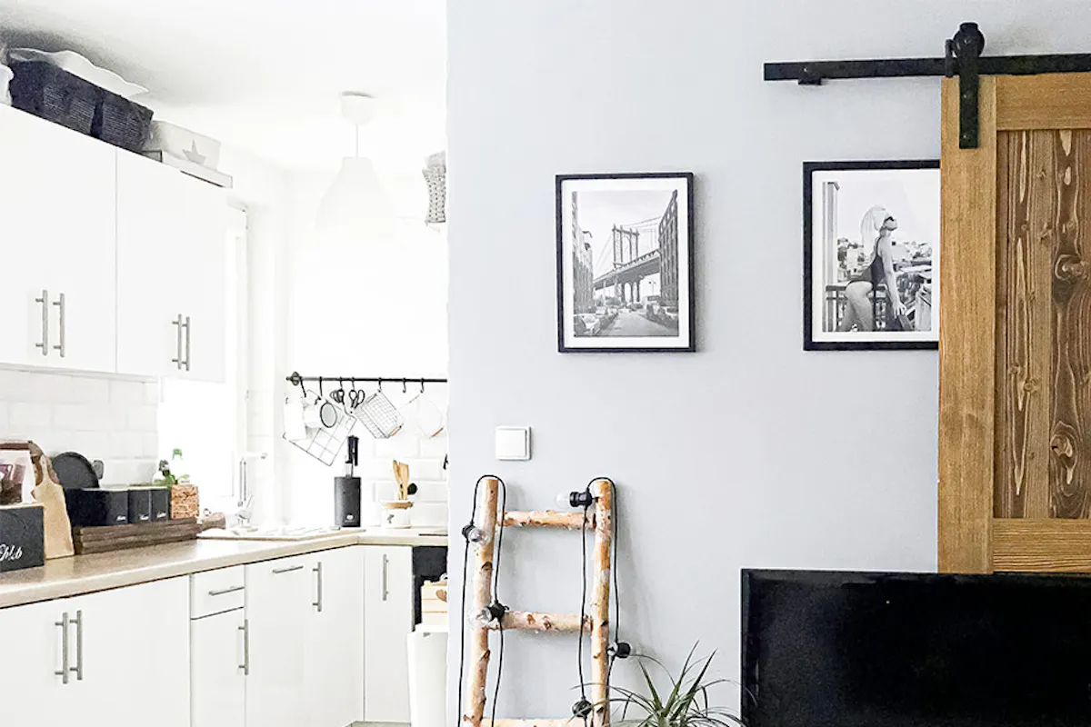 sliding doors in a small apartment, white kitchen with wooden and black accessories, wooden door on black sliding system, metal and wood TV cabinet, black and white posters on the wall, metal and wood decorative elements in the living room