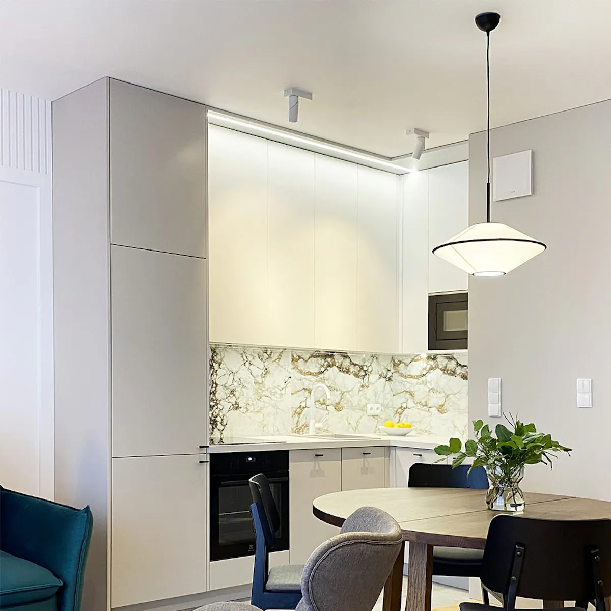 Round walnut dining table with black chairs, light gray kitchen cabinets, decorative stone-pattern tiles above countertop, kitchen row combined with dining area in modern apartment