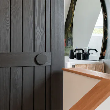 Gros plan sur une porte coulissante en bois noir, dont le grain du bois est visible, dotée d'une poignée ronde noire, avec en arrière-plan l'intérieur d'une salle de bains.