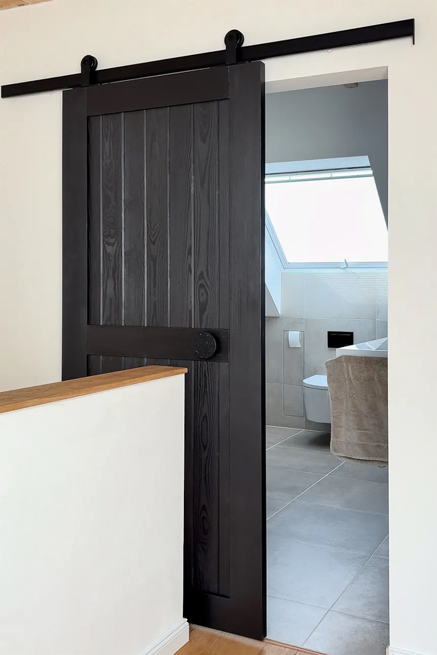 Une porte coulissante noire ouverte, dotée de lamelles verticales et d'une poignée ronde, donnant sur une salle de bains lumineuse équipée d'une lucarne.