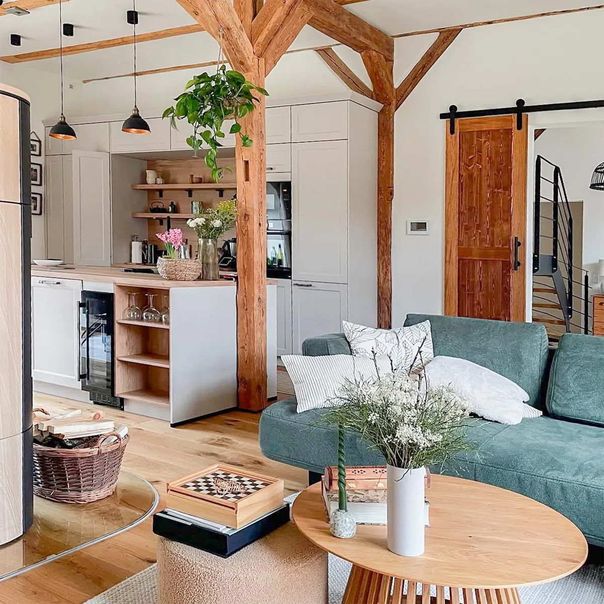 Open-space living room and kitchen in a restored barn, with exposed wooden beams, a green sofa, modern white kitchen units and rustic details.