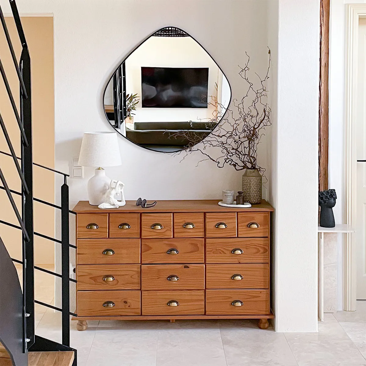 Commode rustique avec de nombreux tiroirs et poignées en laiton, sous un miroir moderne de forme irrégulière, dans un hall lumineux.