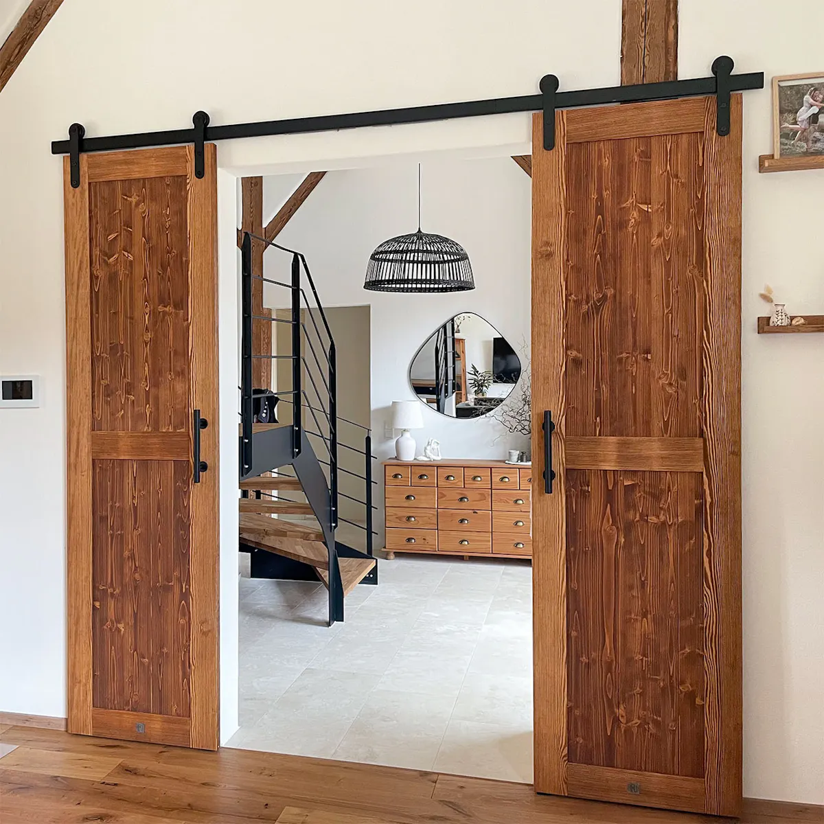 Les doubles portes coulissantes rustiques en bois avec ferrures noires s'ouvrent sur un hall spacieux avec escalier en bois, miroir et commode.