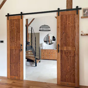 Les doubles portes coulissantes rustiques en bois avec ferrures noires s'ouvrent sur un hall spacieux avec escalier en bois, miroir et commode.