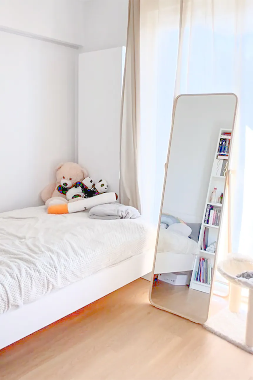Bright bedroom with white bed, stuffed animals, and large standing mirror on wooden floor by window