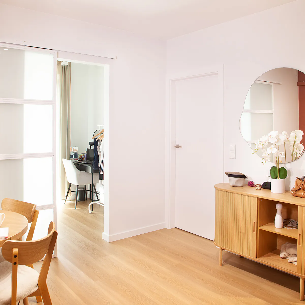 Vue sur l'espace ouvert de la salle à manger et de l'entrée avec une commode en bois et un miroir rond au mur, ainsi qu'une porte coulissante blanche menant à la chambre.