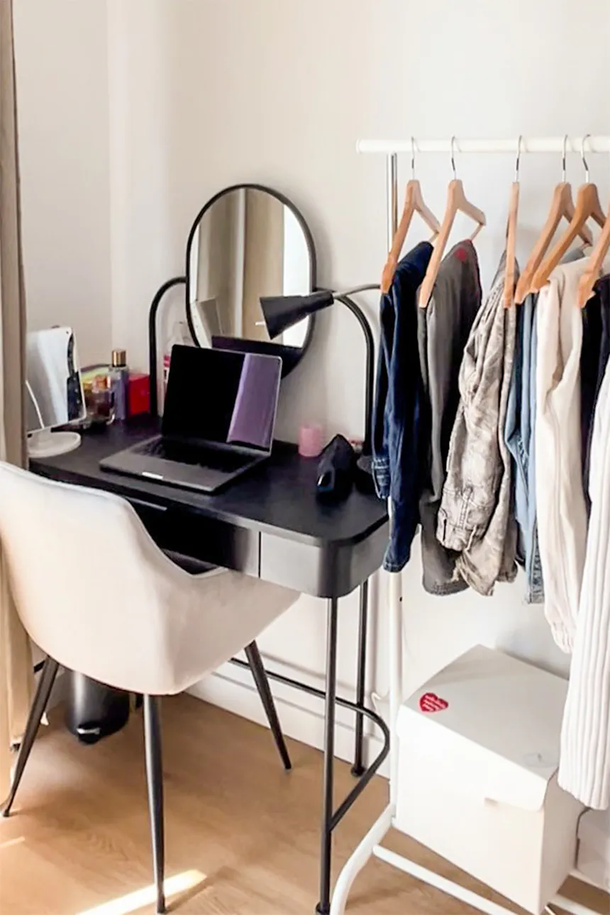 Bureau noir avec un miroir rond, à côté d'une chaise tapissée et d'un portemanteau sur pied dans un intérieur aux murs clairs et au parquet en bois.