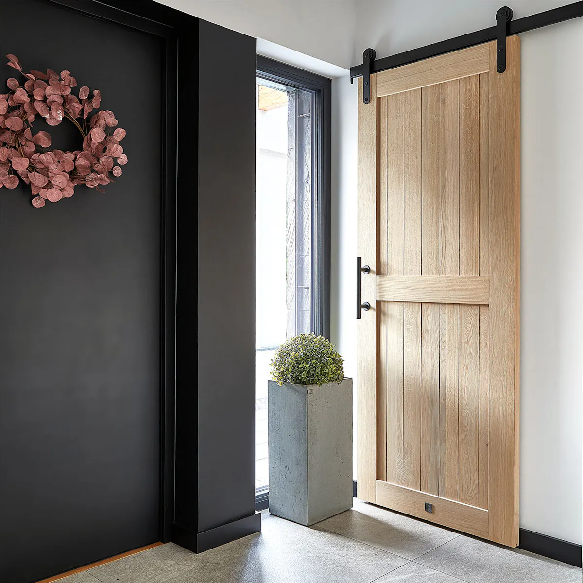A modern vestibule with oak sliding barn doors. Light wooden doors with a black sliding system, alongside a concrete planter and wreath on a dark wall, create an elegant hallway design.