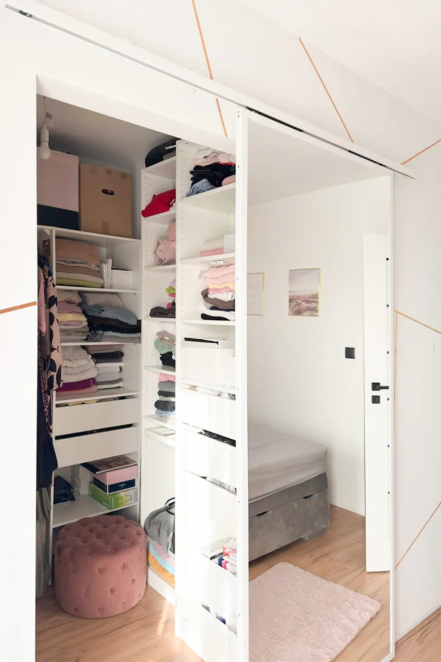 Penderie ouverte blanche avec étagères et tiroirs ; reflet du lit dans le miroir ; pouf en velours couleur poudre dans la penderie.