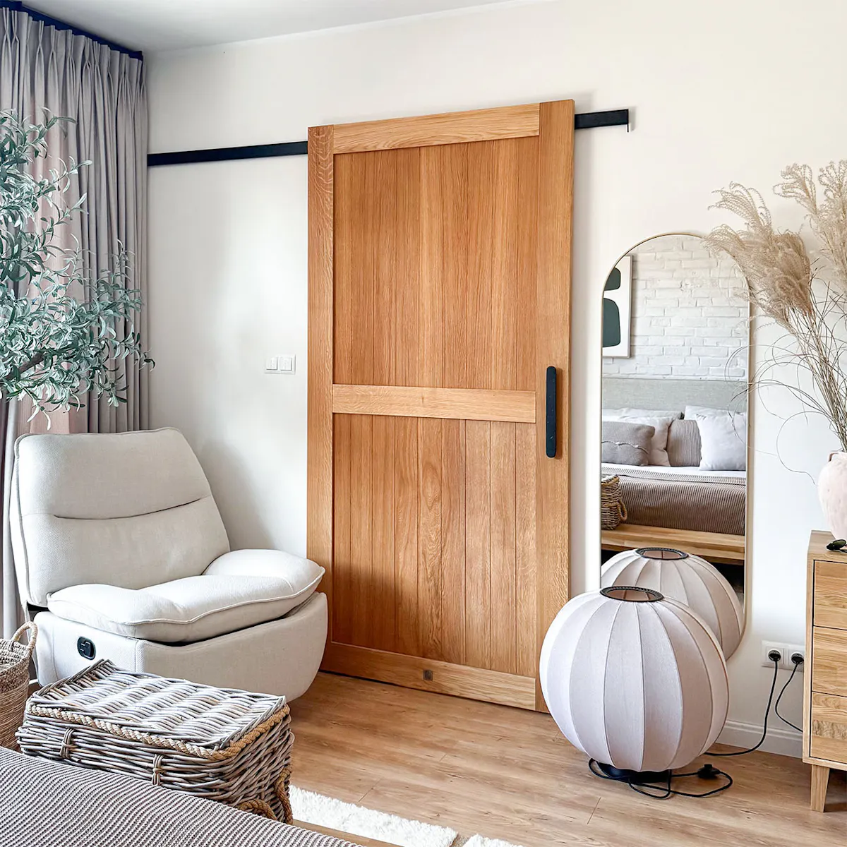 Closed oak sliding doors on a black track in a cosy bedroom. Next to them is a large mirror, woven accessories and light-coloured textiles in the slow living style.