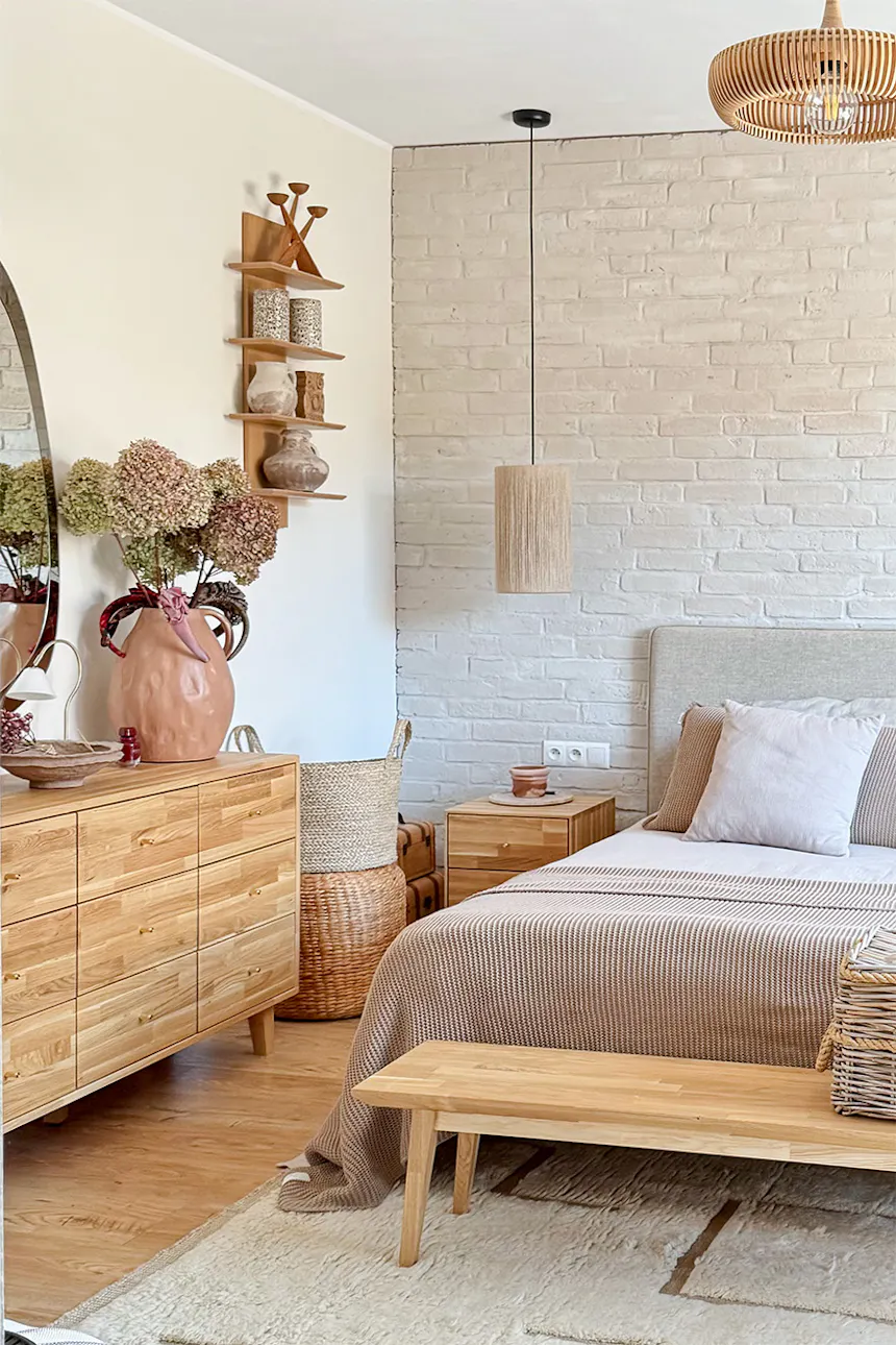 A bedroom design featuring a wooden chest of drawers, a mirror and natural decorations. Visible beige bedspreads, woven baskets and dried plants in a vase.