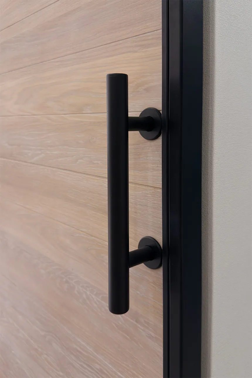 A close-up of a vertical, matt black handle fitted to an oak sliding door panel. The wood’s texture and grain are clearly visible in a bleached oak finish.
