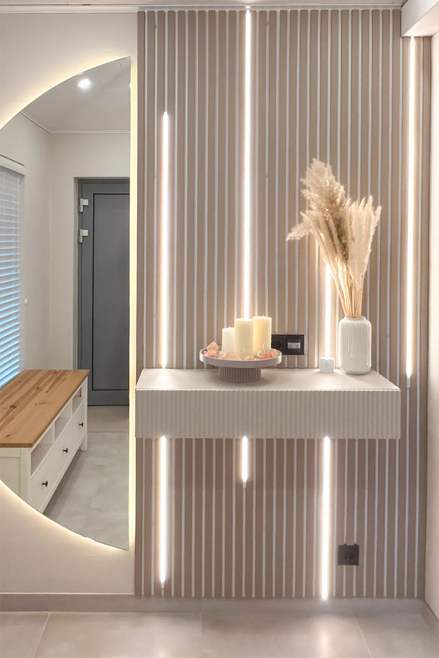 A decorative wall featuring vertical slats and built-in LED strips in a bright hallway. A wall-mounted console table holds decorative candles and a vase of pampas grass. A large, round, backlit mirror hangs on the wall beside it.