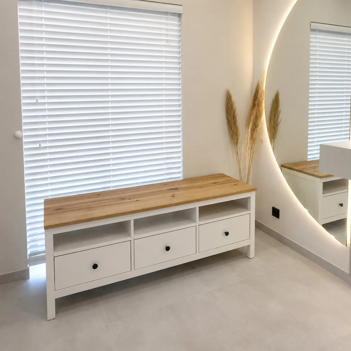A white chest of drawers with a wooden oak top and drawers, standing by a window with white blinds.