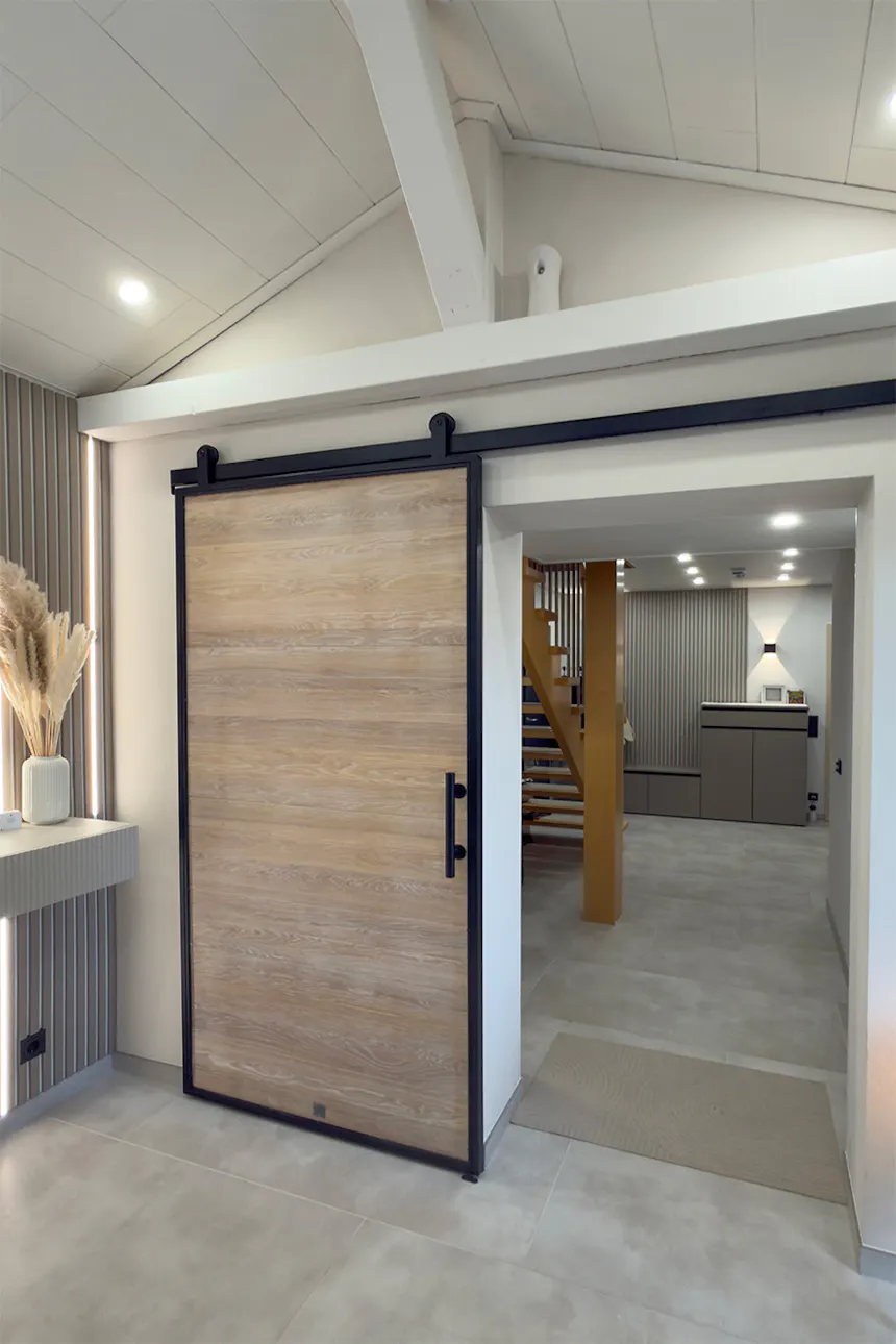 A wide shot of the entrance area with a high, white ceiling and oak sliding doors set in a black loft-style frame. A wooden staircase and hallway can be seen in the background.