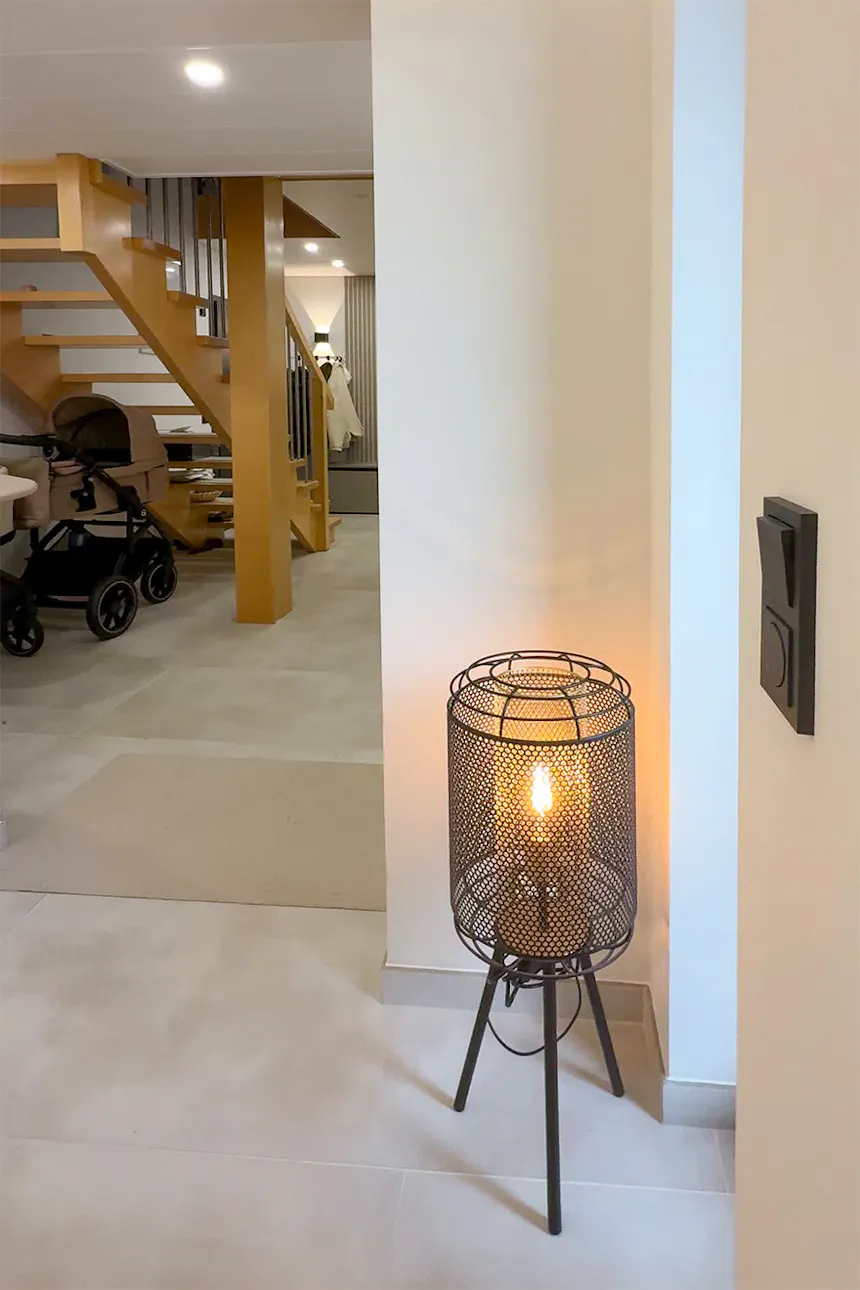 A black, openwork tripod-style metal mesh floor lamp, casting a warm glow in the corner of the room.
