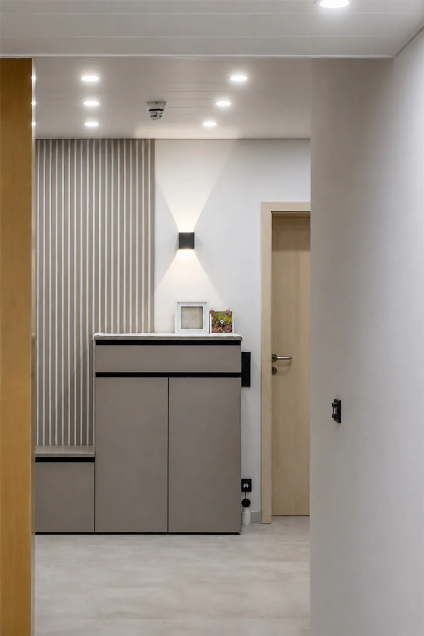 A hallway with minimalist built-in furniture in shades of beige and grey. A modern wall light is mounted on the wall, providing warm, directional lighting.