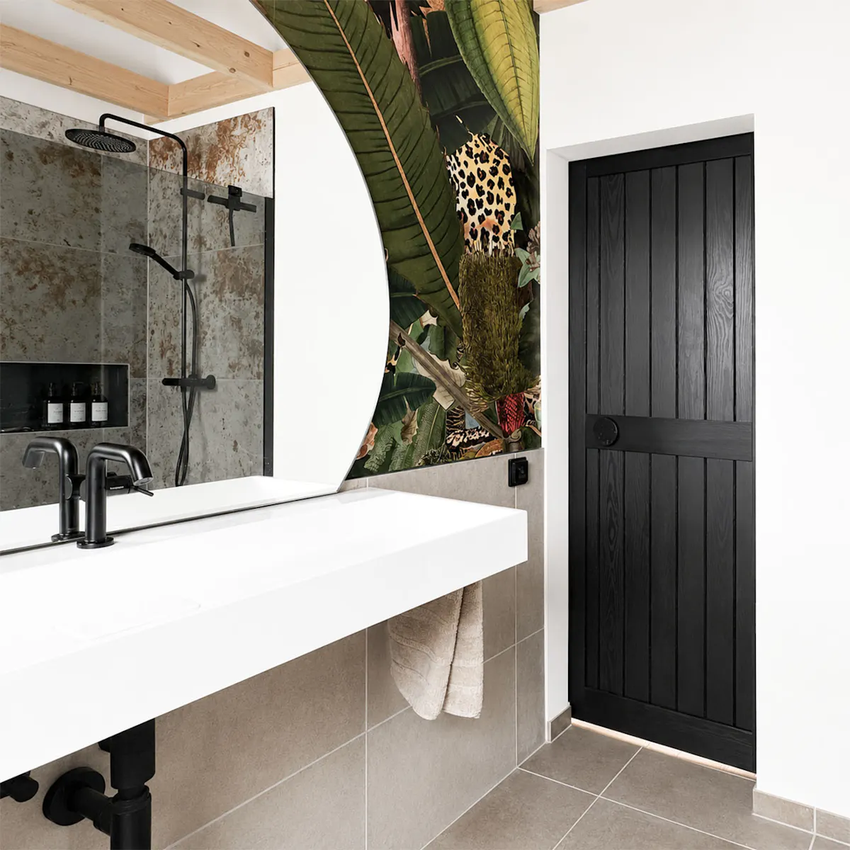Des portes coulissantes en bois noir, vues depuis l'intérieur d'une salle de bains moderne dotée d'un plafond en pente, d'un lavabo blanc et d'un papier peint aux motifs tropicaux.