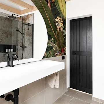 Des portes coulissantes en bois noir, vues depuis l'intérieur d'une salle de bains moderne dotée d'un plafond en pente, d'un lavabo blanc et d'un papier peint aux motifs tropicaux.