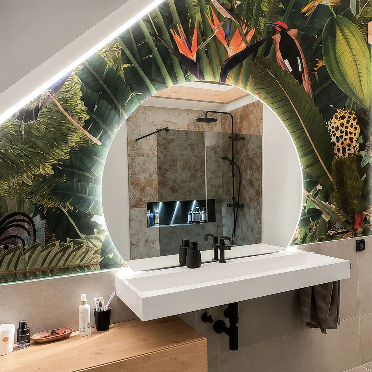 Voici l'espace lavabo de la salle de bains du loft. Un grand miroir rond doté d'un éclairage LED est encastré dans le mur en pente, recouvert d'un papier peint à motifs végétaux.