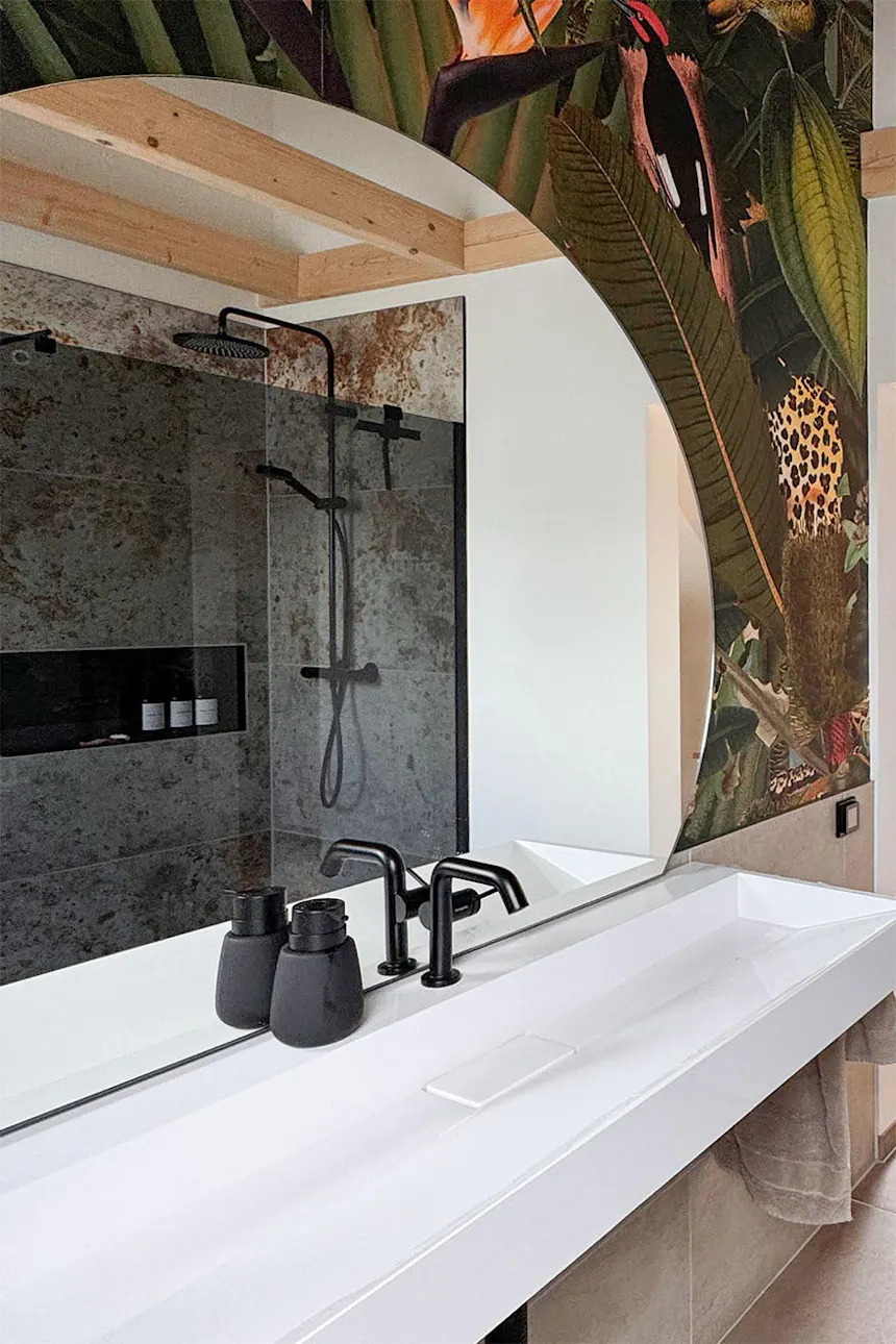 Un gros plan sur la zone du lavabo dans la salle de bains, avec des robinets noirs, le reflet de la cabine de douche dans le miroir et un papier peint aux motifs tropicaux.