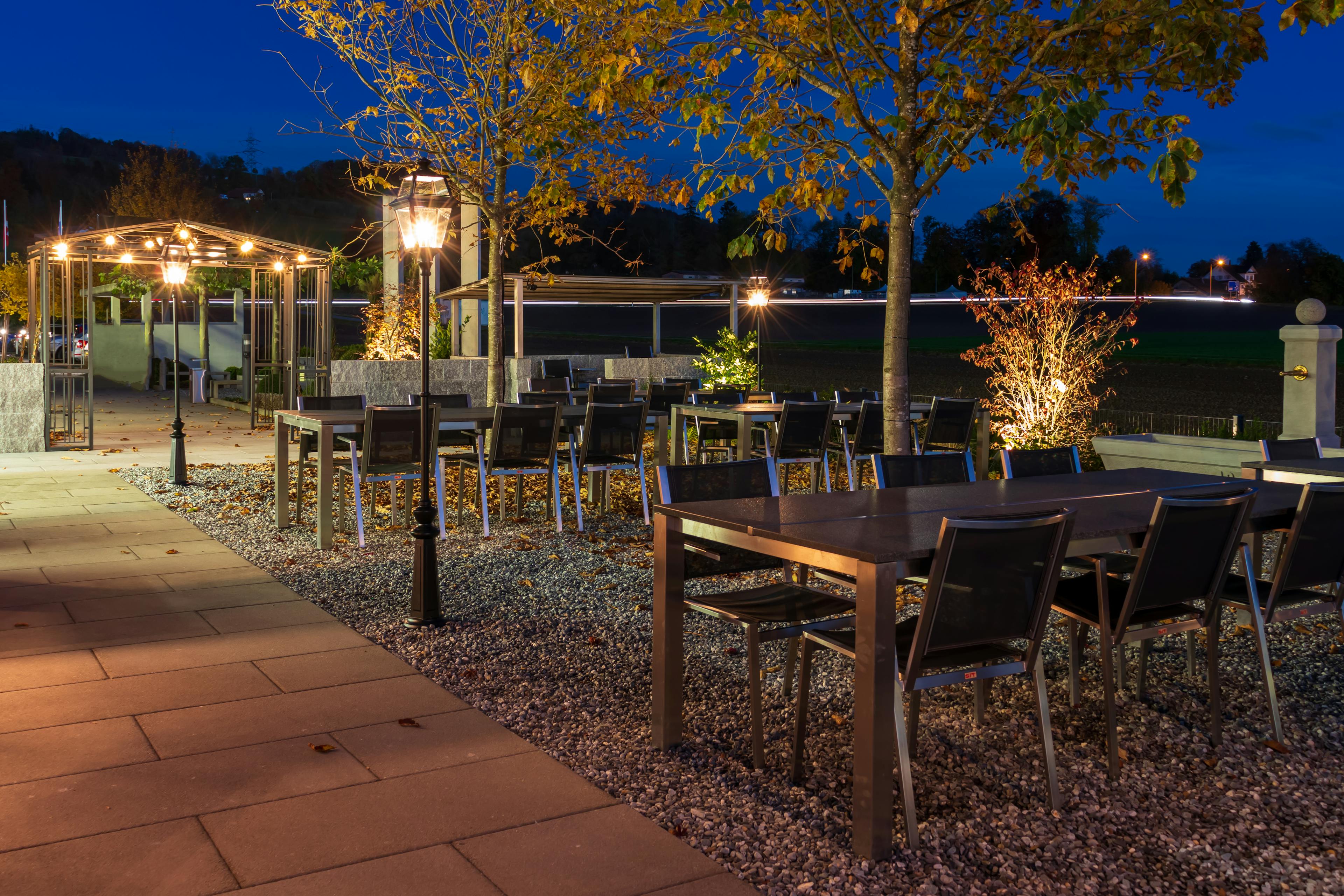 Der Weinereigarten am Abend vorne am Gebäude mit Beleuchtung und Tischgruppen.