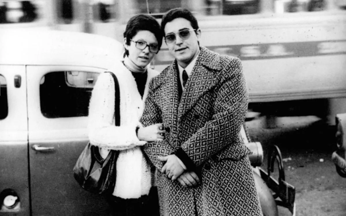 La fotografía en blanco y negro muestra a una pareja —un hombre y una mujer— posando juntos en lo que parece ser un espacio urbano al aire libre. Ambos están de pie muy cerca, con el brazo de la mujer apoyado en el hombro del hombre, transmitiendo cercanía y apoyo mutuo.