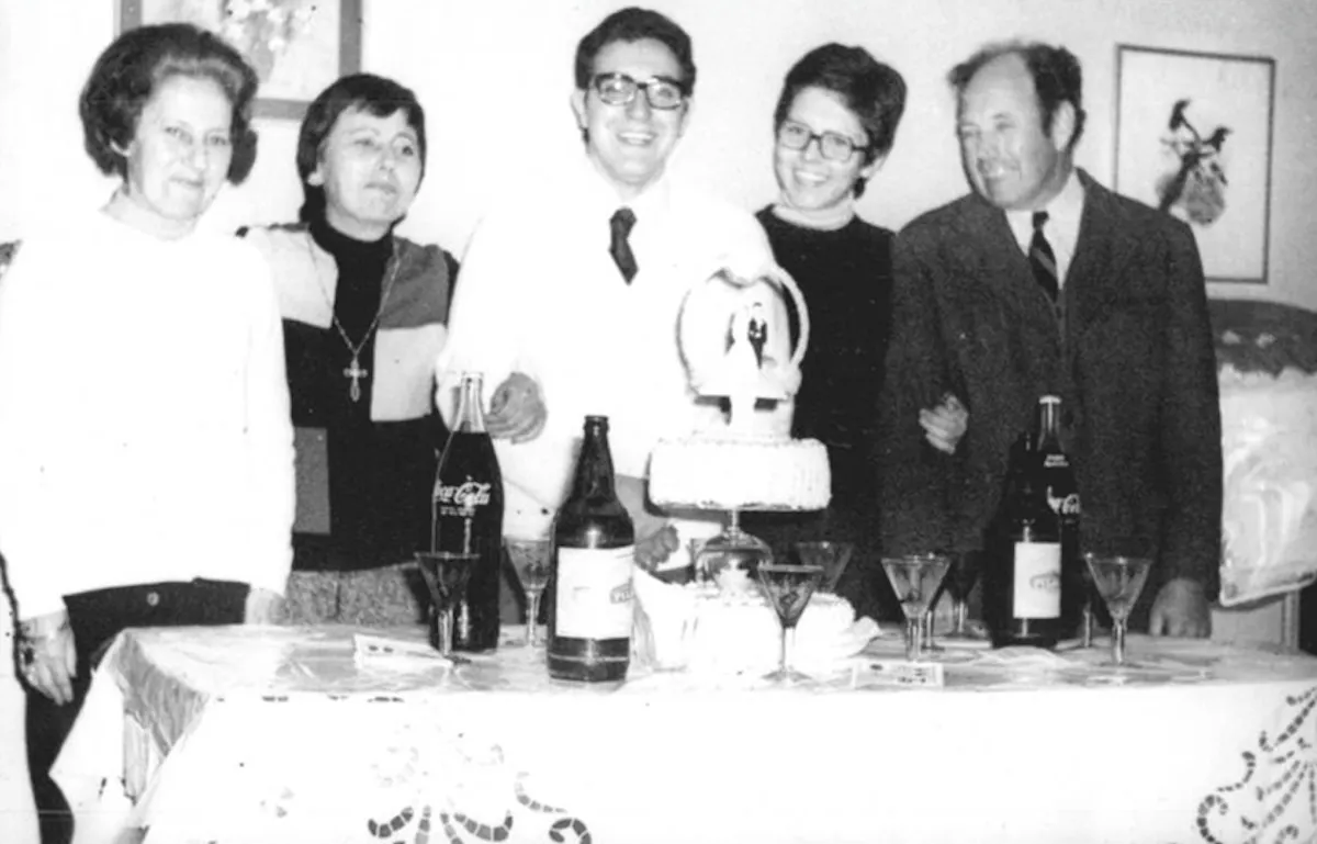 Fotografía en blanco y negro de una escena familiar: el casamiento de Arnone. Cinco personas adultas están de pie detrás de una mesa preparada para una celebración. En el centro hay un hombre joven con gafas, traje claro y corbata oscura. Sonríe ampliamente y parece ser el protagonista del festejo. A su lado, una mujer también sonriente lo toma del brazo.

A la izquierda del joven hay dos mujeres mayores; una viste ropa clara y la otra un conjunto oscuro con un collar largo y una cruz visible sobre el pecho. A la derecha hay un hombre mayor con traje oscuro y corbata, que mira hacia el joven con expresión afectuosa.

Sobre la mesa hay una torta decorada colocada en una base alta; en la parte superior se distingue una figura decorativa. También hay varias botellas y copas distribuidas frente a ellos. El mantel tiene bordados visibles en el borde.