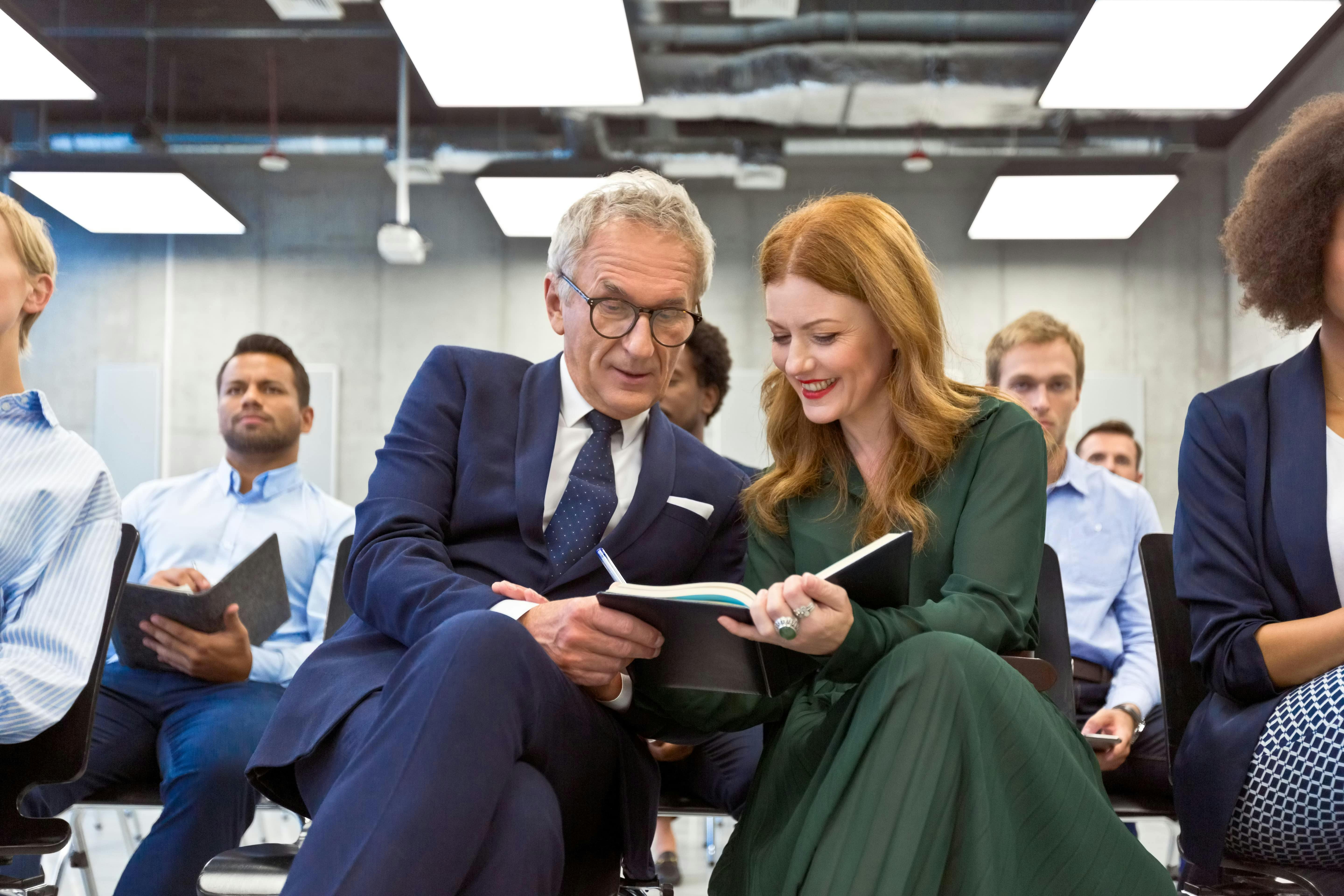A picture of a caucasian man with glasses and a caucasian women with red hair sharing a notebook surrounded by a group of workers 