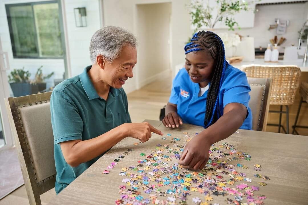 A caregiver helps her elderly client put together a puzzle on the dining room table.