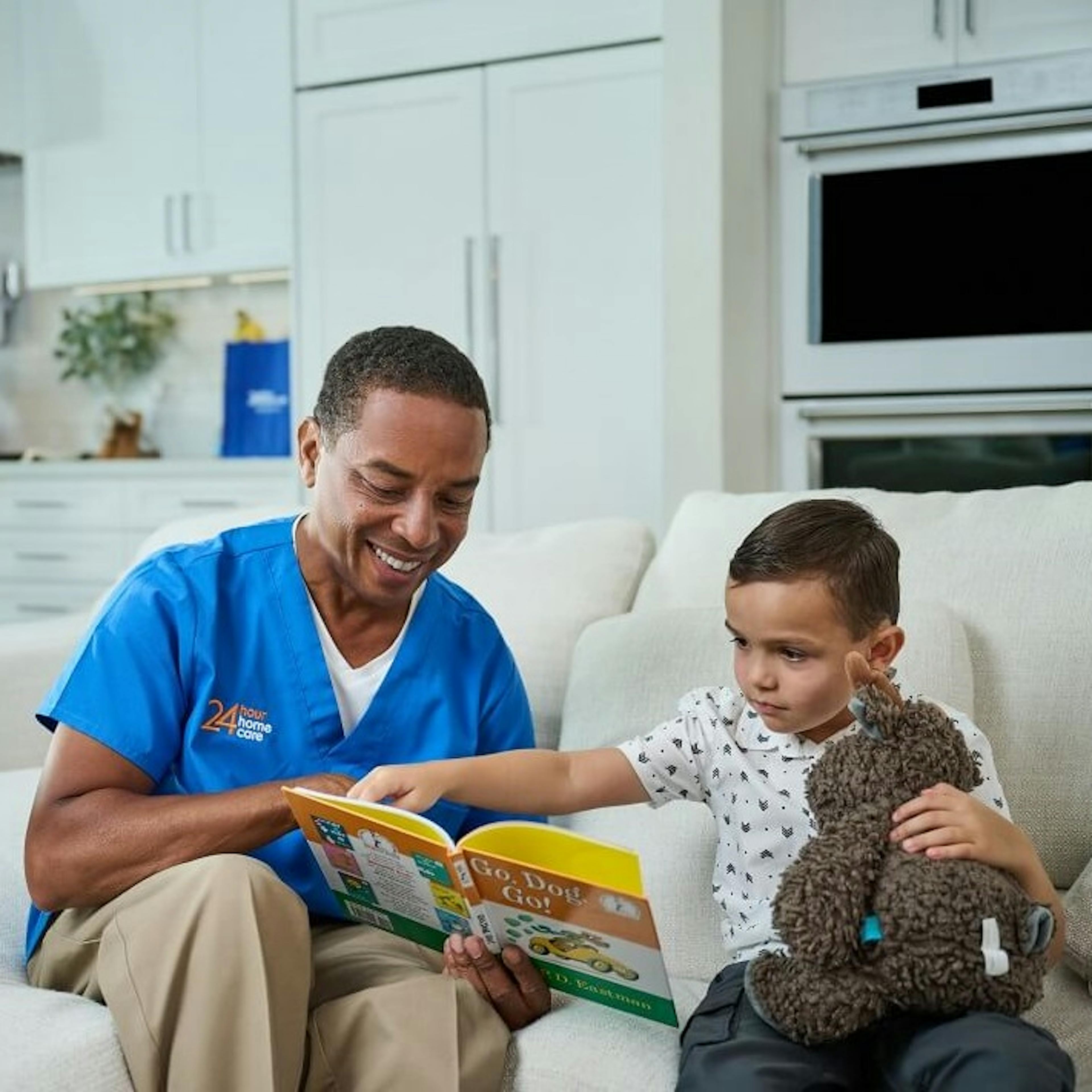 A caregiver reads with his child client on the couch while the mother does dishes in the background.
