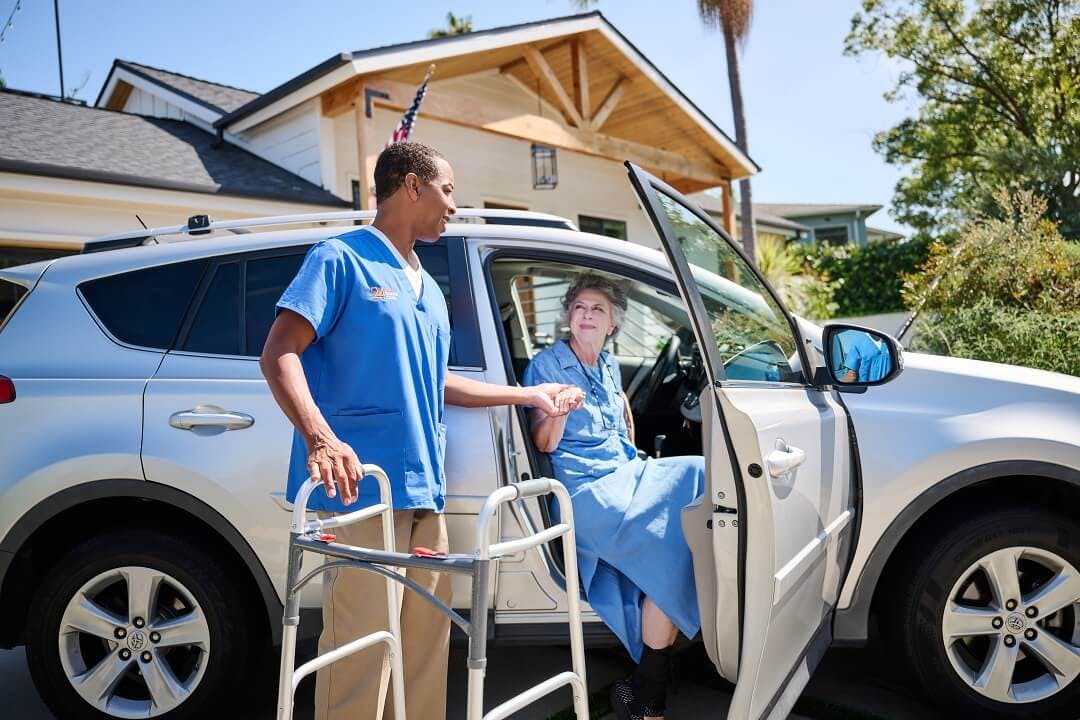 A male caregiver helps an elderly client out of her car and into a walker.
