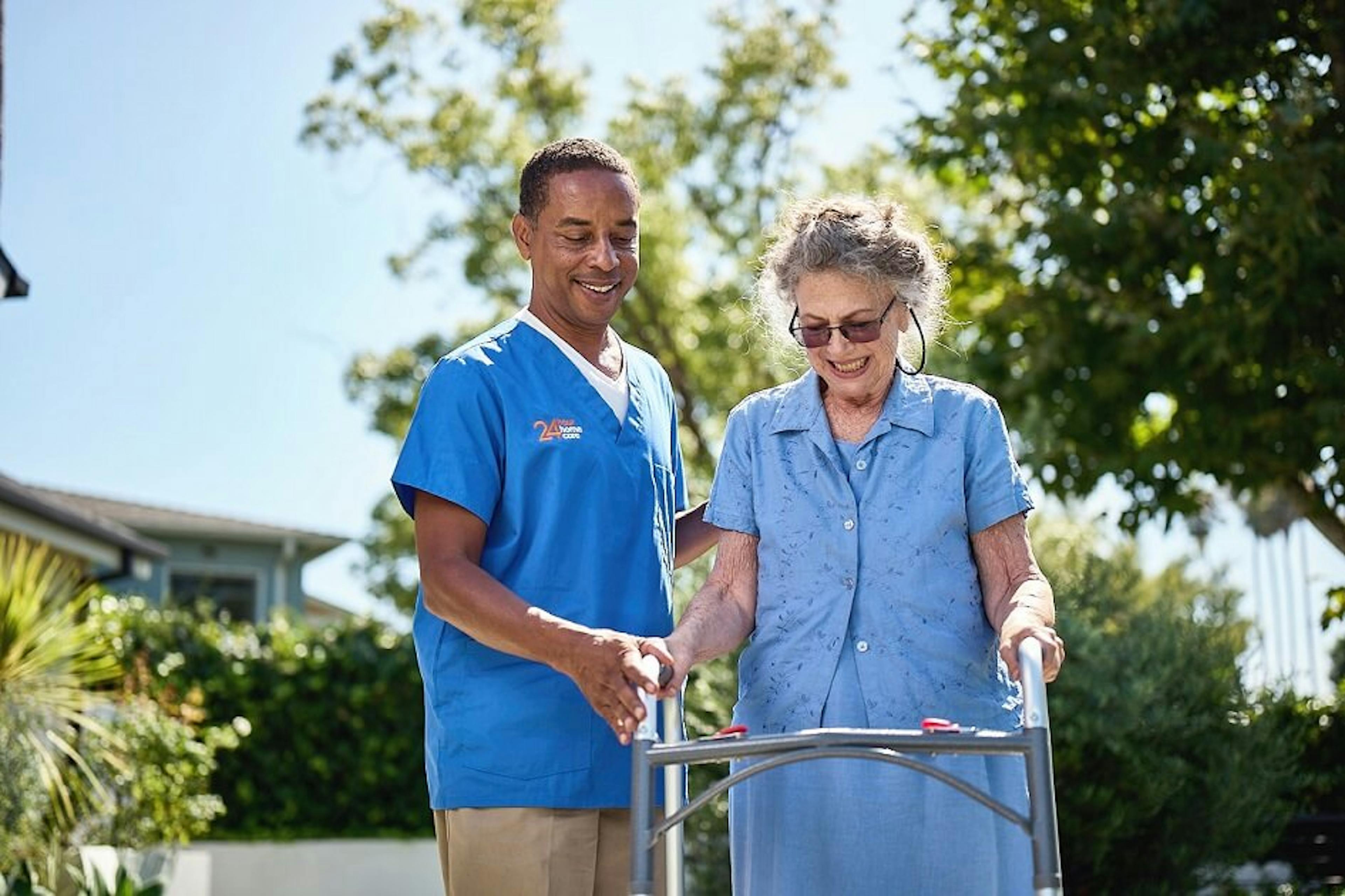 A male caregiver helps his elderly client walk outside with her walker. 