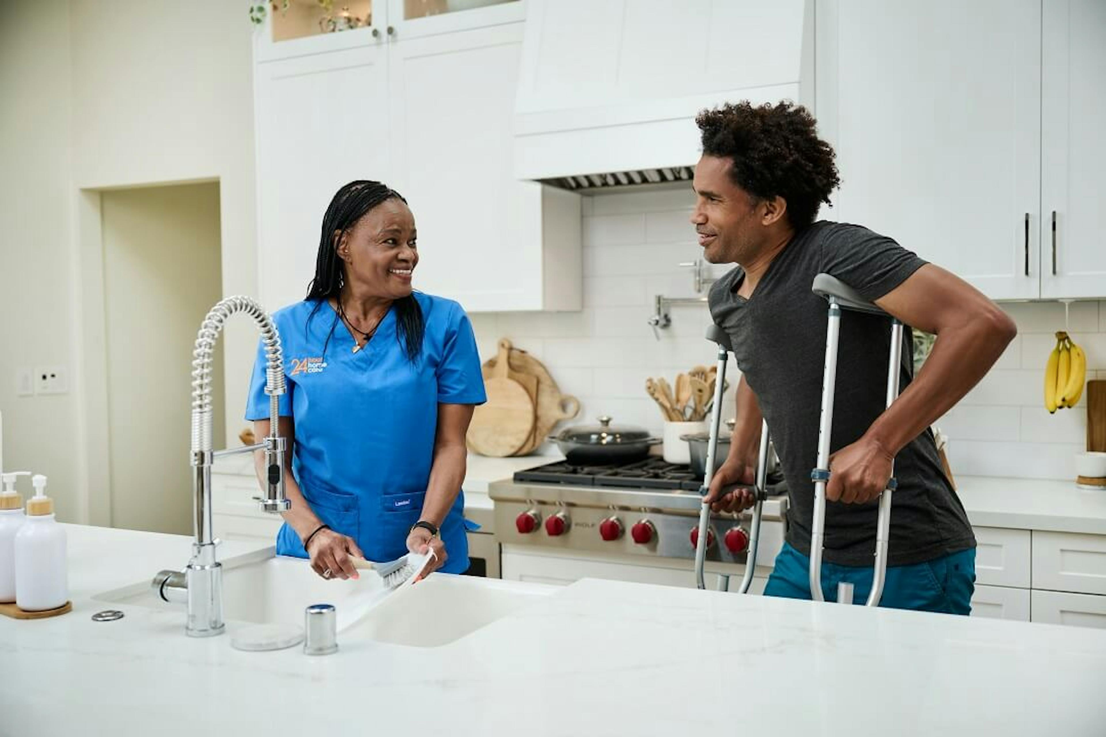 A female caregiver assists an adult client on crutches in the kitchen.