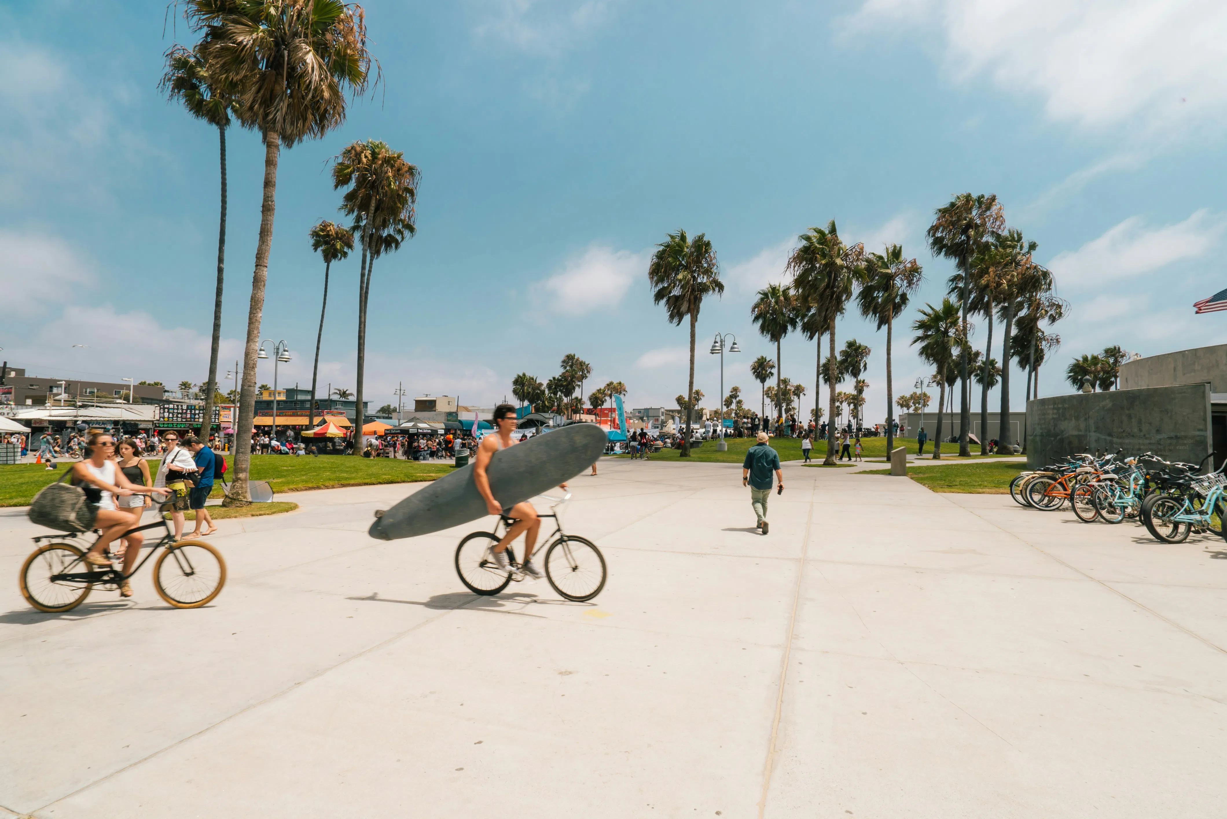 The Venice boardwalk a short walk from the building: palms along Ocean Front Walk, people cycling past, and a surfer carrying a longboard.
