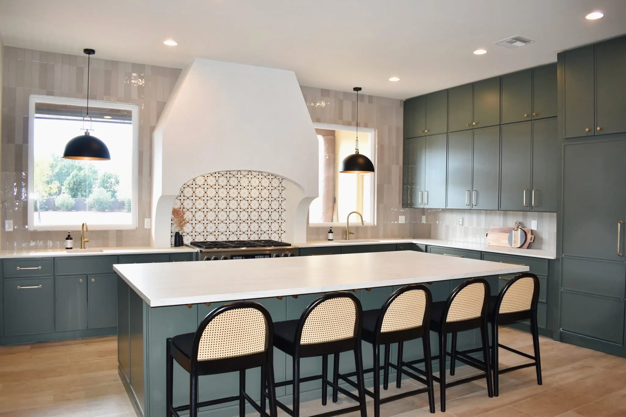 Wide view of green cabinetry, oversized white kitchen island, brass hardware, and black pendant lights.