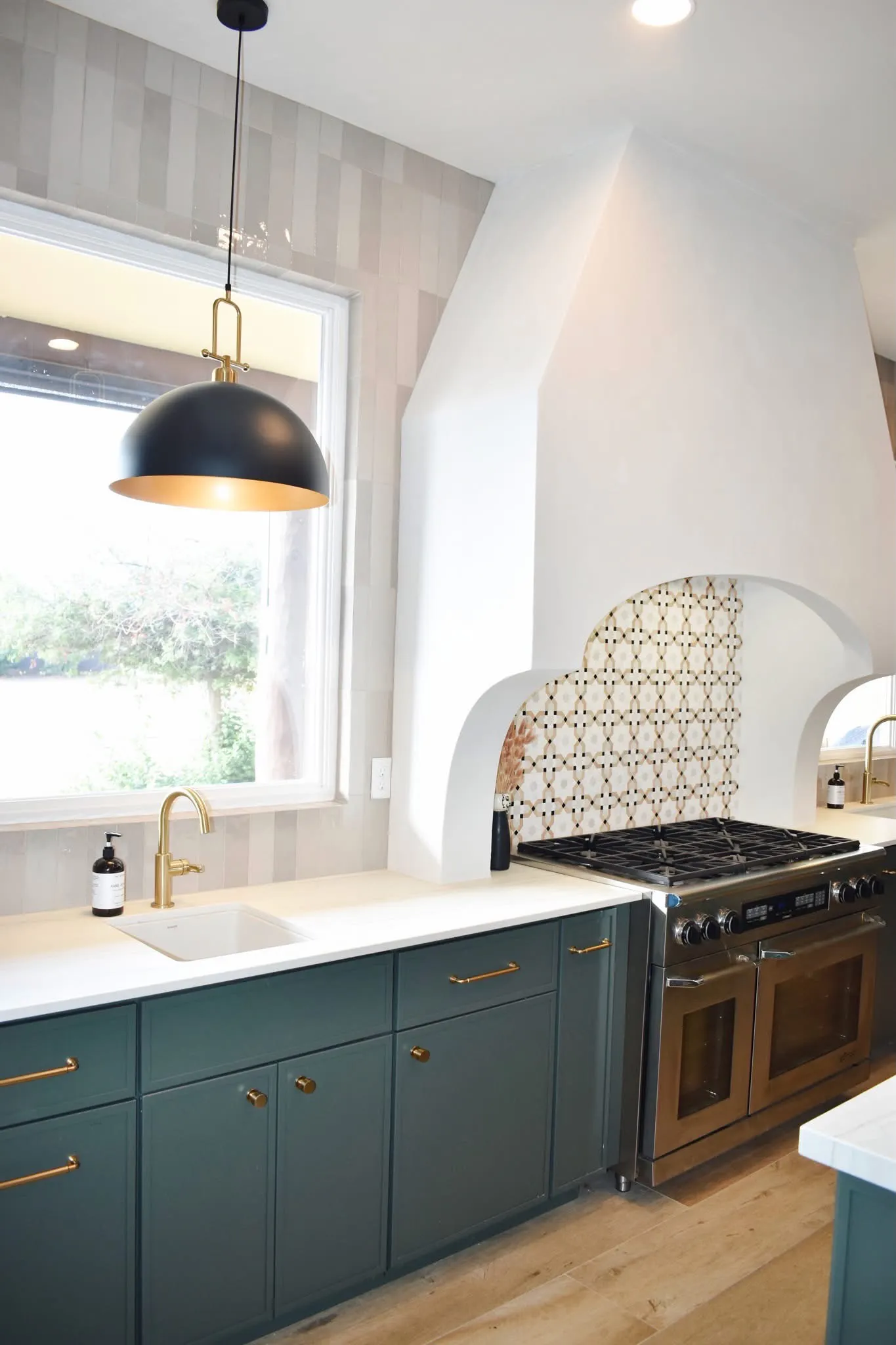 Green kitchen cabinetry beside a stainless range, sculptural plaster hood, patterned tile, and brass pot filler.