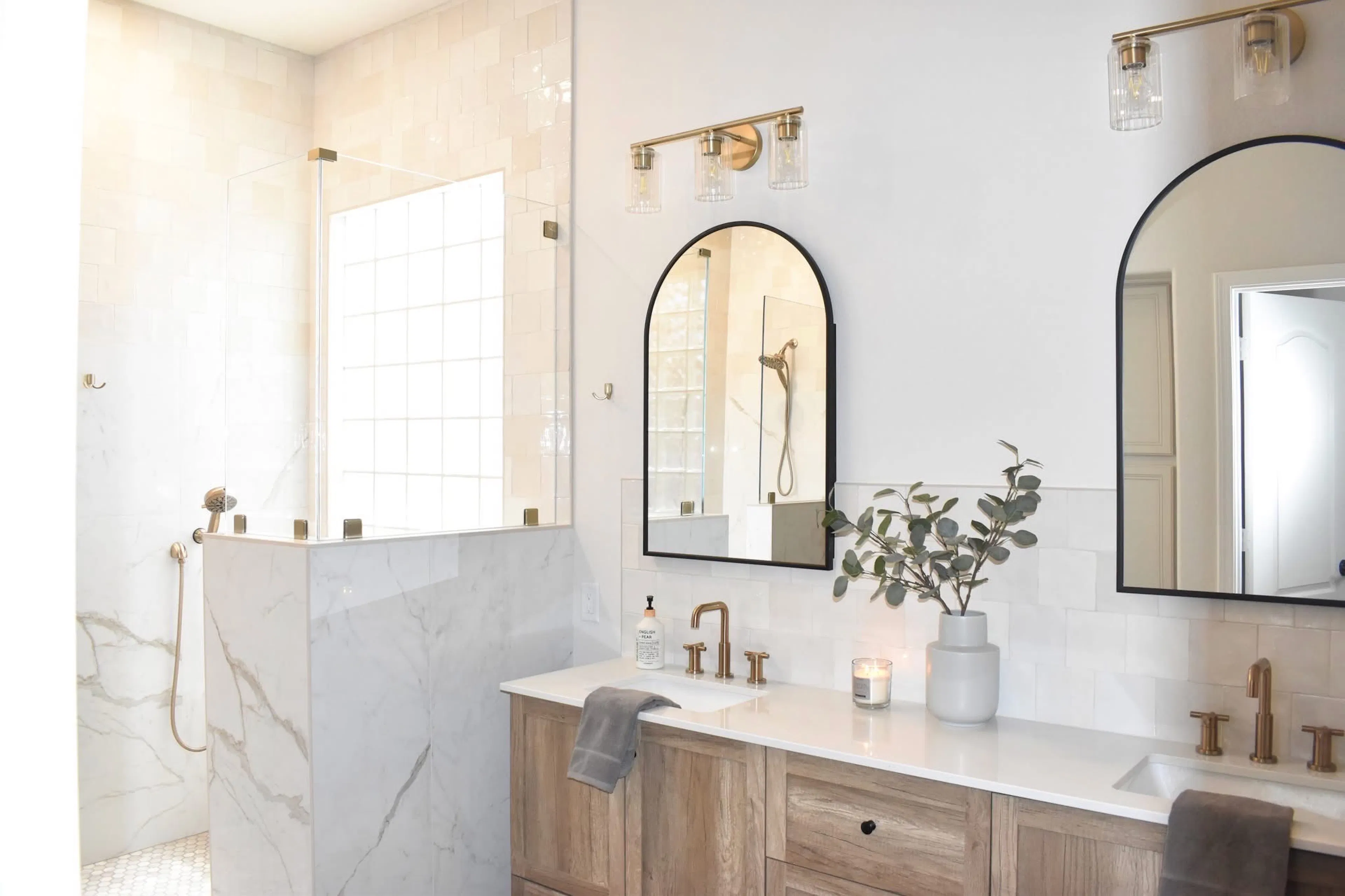 Remodeled bathroom with a glass shower, marble-look tile, wood vanity, arched mirrors, and brass fixtures.
