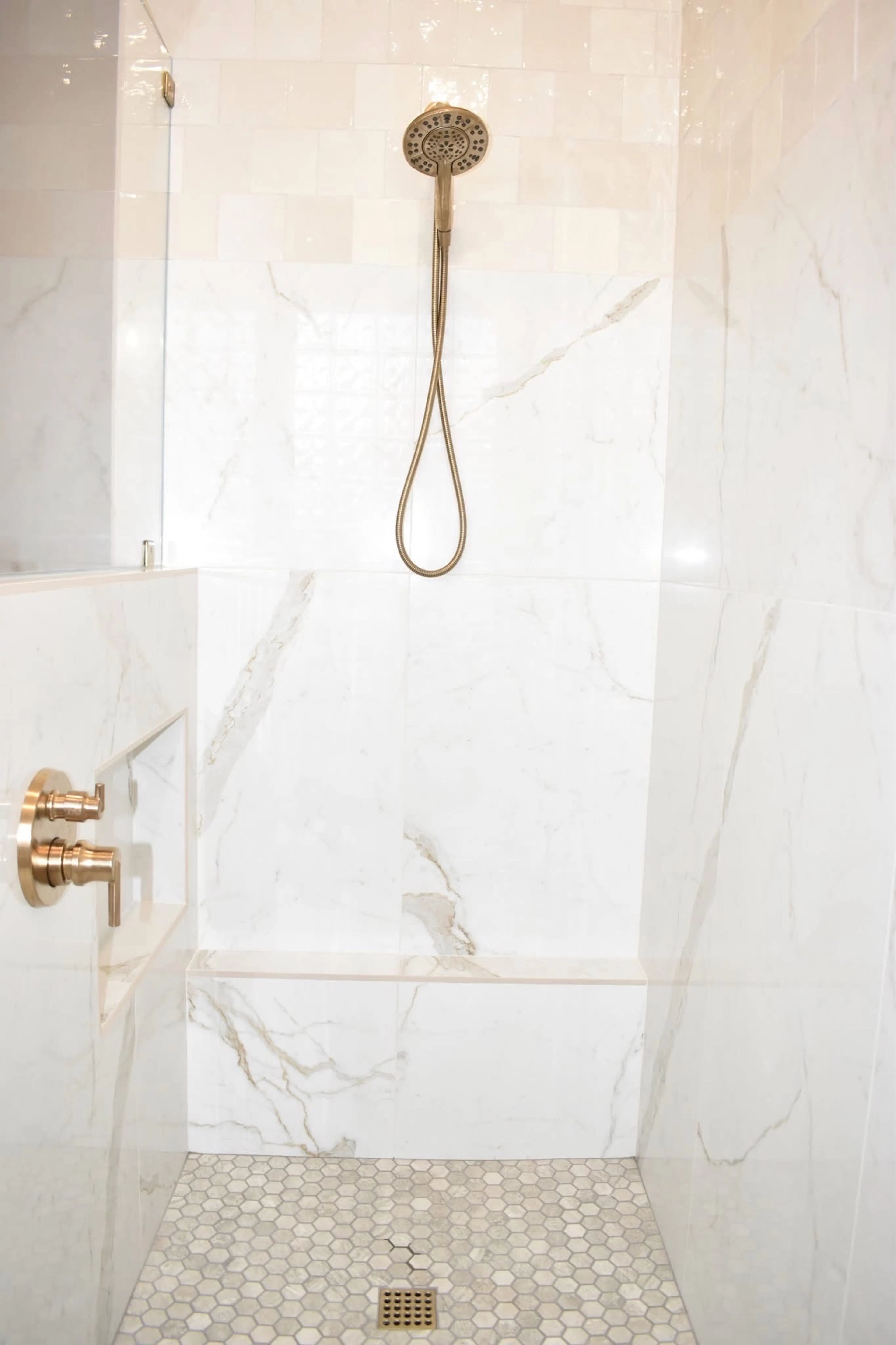 Walk-in shower with marble-look wall panels, glossy ivory tile, brass hand shower, bench, and mosaic floor.